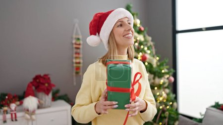 Téléchargez les photos : Jeune femme blonde tenant cadeau debout près de l'arbre de Noël à la maison - en image libre de droit