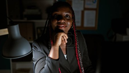 Téléchargez les photos : Femme d'affaires afro-américaine confiante réfléchissant à des idées, travaillant tard au bureau, éclairée par la lumière de l'écran de l'ordinateur - en image libre de droit