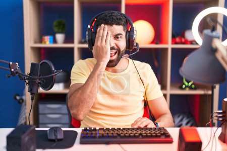 Téléchargez les photos : Homme hispanique avec barbe jouant à des jeux vidéo avec écouteurs couvrant un oeil avec la main, sourire confiant sur le visage et l'émotion surprise. - en image libre de droit