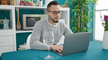 Photo for Hispanic man drinking glass of wine sitting on table using laptop at dinning room - Royalty Free Image