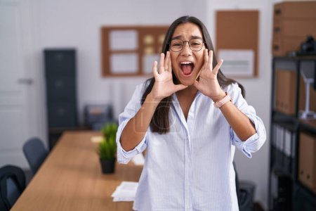 Foto de Joven mujer hispana en la oficina gritando furiosa con las manos sobre la boca - Imagen libre de derechos