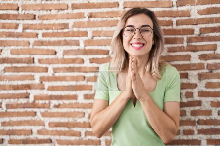 Photo for Young beautiful woman standing over bricks wall praying with hands together asking for forgiveness smiling confident. - Royalty Free Image