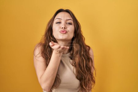 Téléchargez les photos : Jeune femme hispanique debout sur fond jaune regardant la caméra souffler un baiser avec la main sur l'air étant belle et sexy. expression d'amour. - en image libre de droit