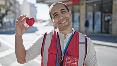 Photo for Young hispanic man volunteer smiling holding red heart for blood donation at street - Royalty Free Image