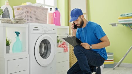 Photo for Young hispanic man technician repairing washing machine talking on smartphone at laundry room - Royalty Free Image