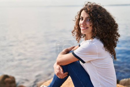 Téléchargez les photos : Jeune femme hispanique souriante confiante assise sur le rocher au bord de la mer - en image libre de droit