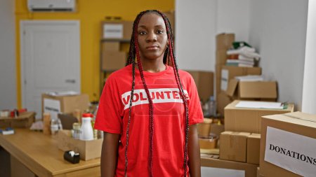 Photo for Beautiful african american woman, a serious-faced volunteer, stands indoors in charity center's storehouse, adult worker in t-shirt, unified in altruism behind packed donations - Royalty Free Image