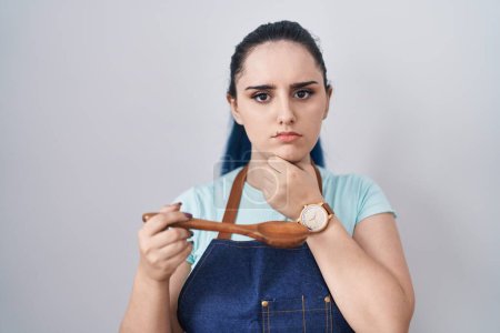 Téléchargez les photos : Jeune fille moderne avec les cheveux bleus portant tablier de cuisine tenant cuillère visage sérieux penser à la question avec la main sur le menton, réfléchi sur l'idée déroutante - en image libre de droit
