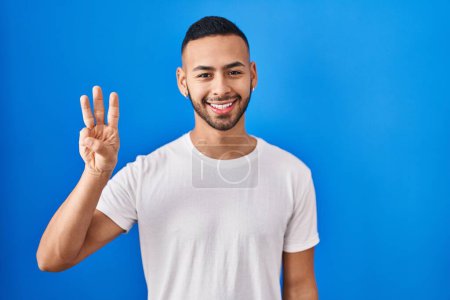 Photo for Young hispanic man standing over blue background showing and pointing up with fingers number three while smiling confident and happy. - Royalty Free Image