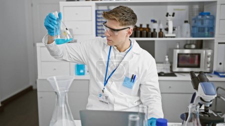 Téléchargez les photos : Jeune homme caucasien scientifique mesurant le liquide sur éprouvette à l'aide d'un ordinateur portable au laboratoire - en image libre de droit