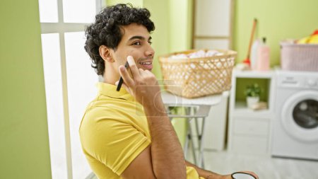 Téléchargez les photos : Jeune homme latin écoutant un message vocal par smartphone boire du café en attendant la machine à laver à la buanderie - en image libre de droit