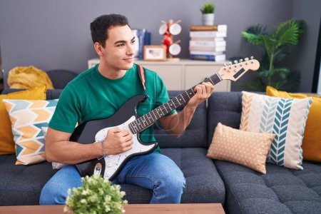 Téléchargez les photos : Jeune homme hispanique jouant de la guitare électrique assis sur le canapé à la maison - en image libre de droit