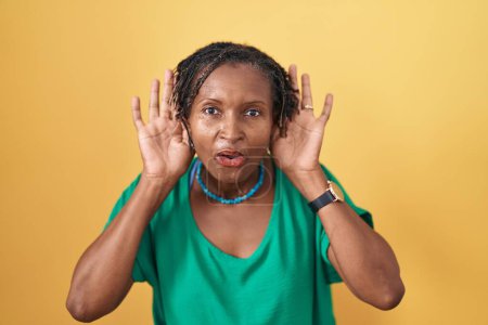 Foto de Mujer africana con rastas de pie sobre fondo amarillo tratando de escuchar ambas manos en el gesto de la oreja, curiosa de chismes. problema auditivo, sordo - Imagen libre de derechos