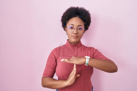 Téléchargez les photos : Belle femme africaine avec les cheveux bouclés debout sur fond rose faisant un geste de temps avec les mains, frustré et sérieux visage - en image libre de droit