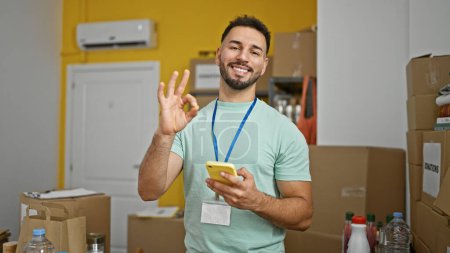 Téléchargez les photos : Jeune homme arabe bénévole en utilisant smartphone faire un geste correct au centre de charité - en image libre de droit