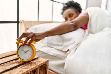Foto de Mujer afroamericana apagando el despertador tumbado en la cama en el dormitorio - Imagen libre de derechos