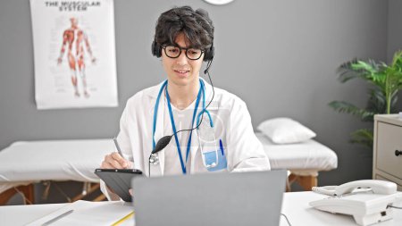 Foto de Joven médico hispano haciendo videollamada con computadora portátil escribiendo en touchpad en la clínica - Imagen libre de derechos