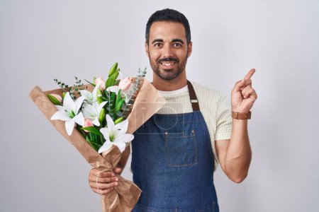 Téléchargez les photos : Homme hispanique avec barbe travaillant comme fleuriste avec un grand sourire sur le visage, pointant du doigt vers le côté en regardant la caméra. - en image libre de droit