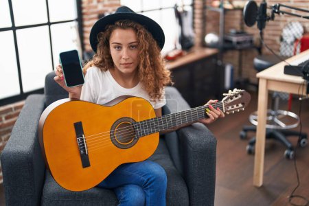 Foto de Mujer caucásica joven tocando la guitarra clásica en el estudio de música sosteniendo teléfono inteligente expresión despistada y confusa. concepto de duda. - Imagen libre de derechos