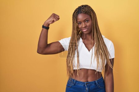 Foto de Mujer afroamericana con cabello trenzado de pie sobre fondo amarillo persona fuerte mostrando el músculo del brazo, confiado y orgulloso de poder - Imagen libre de derechos