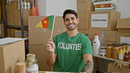 Foto de Joven hombre hispano voluntario sonriendo confiado sosteniendo bandera de camerún en el centro de caridad - Imagen libre de derechos