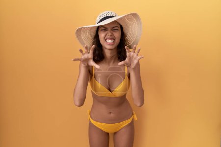Téléchargez les photos : Jeune femme hispanique portant un bikini et un chapeau d'été souriant drôle faisant geste griffe comme chat, expression agressive et sexy - en image libre de droit