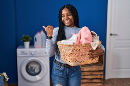 Photo for African american woman holding laundry basket pointing thumb up to the side smiling happy with open mouth - Royalty Free Image