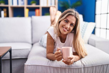 Téléchargez les photos : Jeune femme blonde buvant du café allongé sur le canapé à la maison - en image libre de droit
