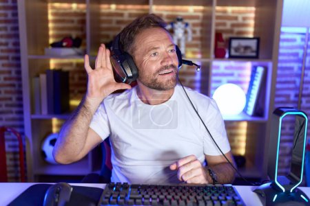 Téléchargez les photos : Homme d'âge moyen avec barbe jouant à des jeux vidéo portant un casque souriant avec la main sur l'oreille en écoutant une rumeur ou des commérages. concept de surdité. - en image libre de droit