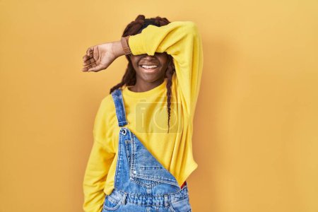 Photo for African woman standing over yellow background covering eyes with arm smiling cheerful and funny. blind concept. - Royalty Free Image