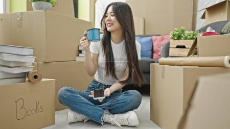 Photo for Young chinese woman using smartphone drinking coffee sitting on floor at new home - Royalty Free Image