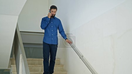 Foto de Un joven trabajador de negocios hispano hablando por teléfono inteligente bajando las escaleras de la oficina - Imagen libre de derechos