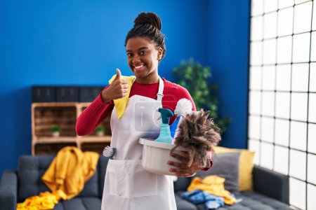 Téléchargez les photos : Jeune afro-américain avec des tresses de nettoyage de la maison tenant des produits de nettoyage souriant heureux et positif, pouce levé faire excellent et signe d'approbation - en image libre de droit