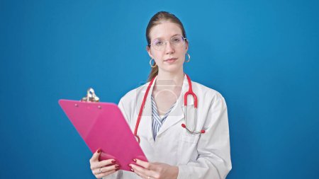 Foto de Joven mujer rubia médico leyendo documento en portapapeles diciendo no con la cabeza sobre fondo azul aislado - Imagen libre de derechos