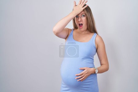 Téléchargez les photos : Jeune femme enceinte debout sur fond blanc surpris avec la main sur la tête pour erreur, rappelez-vous erreur. oublié, mauvais concept de mémoire. - en image libre de droit