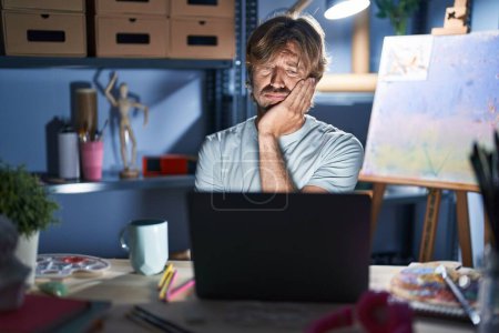 Téléchargez les photos : Homme d'âge moyen assis au studio d'art avec ordinateur portable la nuit pensant fatigué et ennuyé avec des problèmes de dépression avec les bras croisés. - en image libre de droit