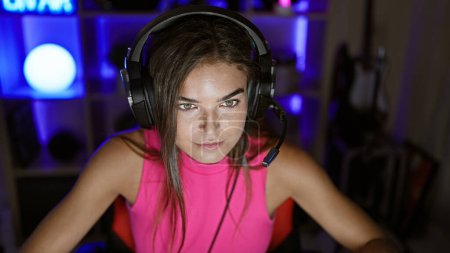 Foto de Atractiva joven streamer hispana, atrapada en la noche cibernética, totalmente concentrada y con auriculares mientras juega un videojuego de alto riesgo en su oscura sala de juegos en casa - Imagen libre de derechos