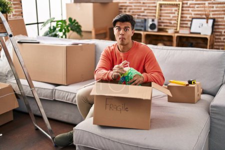 Photo for Hispanic man moving to a new home unpacking making fish face with mouth and squinting eyes, crazy and comical. - Royalty Free Image