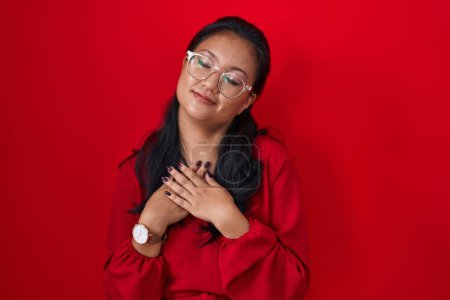 Téléchargez les photos : Asiatique jeune femme debout sur fond rouge souriant avec les mains sur la poitrine avec les yeux fermés et geste reconnaissant sur le visage. concept de santé. - en image libre de droit