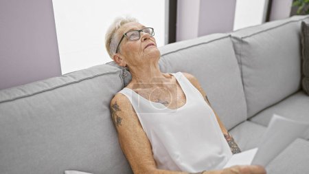 Téléchargez les photos : Femme âgée aux cheveux gris trouve le confort dans la détente sur un canapé confortable, tenant du papier à l'intérieur dans son salon ensoleillé, présentant une expression sérieuse - en image libre de droit