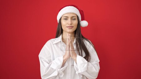 Téléchargez les photos : Jeune belle femme hispanique souriante priant portant un chapeau de Noël sur fond rouge isolé - en image libre de droit