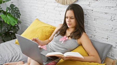 Photo for Young beautiful hispanic woman using laptop holding notebook sitting on bed at bedroom - Royalty Free Image