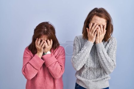 Téléchargez les photos : Mère et fille debout sur fond bleu avec une expression triste couvrant le visage des mains tout en pleurant. concept de dépression. - en image libre de droit