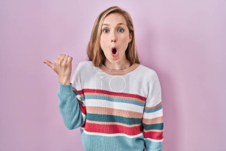 Photo for Young blonde woman standing over pink background surprised pointing with hand finger to the side, open mouth amazed expression. - Royalty Free Image