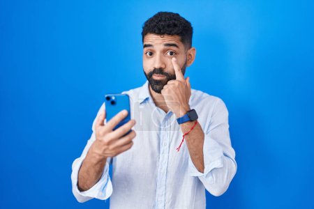 Téléchargez les photos : Homme hispanique avec barbe en utilisant un message de dactylographie smartphone pointant vers l'œil vous regardant geste, expression suspecte - en image libre de droit