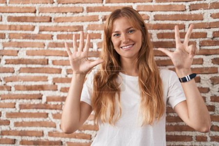 Foto de Joven mujer caucásica de pie sobre la pared de ladrillos mostrando y señalando hacia arriba con los dedos número ocho mientras sonríe confiado y feliz. - Imagen libre de derechos