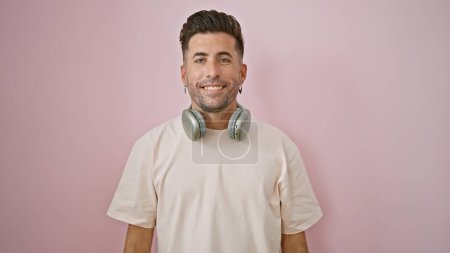 Foto de Sonriente joven hispano disfrutando de la música, auriculares balanceándose de pie sobre un fondo rosa aislado - fresco, confiado y casualmente guapo - Imagen libre de derechos