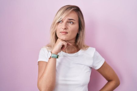 Foto de Young blonde woman standing over pink background with hand on chin thinking about question, pensive expression. smiling with thoughtful face. doubt concept. - Imagen libre de derechos