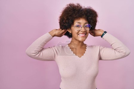 Foto de Joven mujer afroamericana de pie sobre fondo rosa sonriendo tirando de las orejas con los dedos, gesto divertido. problema de audición - Imagen libre de derechos