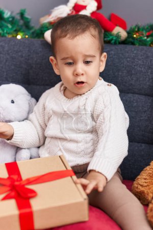 Photo for Adorable hispanic toddler unpacking christmas gift sitting on sofa at home - Royalty Free Image